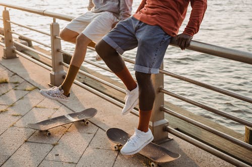 Crop anonymous multiracial males skaters wearing casual summer outfits sitting together on embankment railing in evening sun