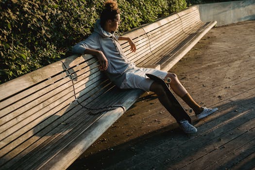 Young man relaxing on a bench with a skateboard in a sunlit urban park.