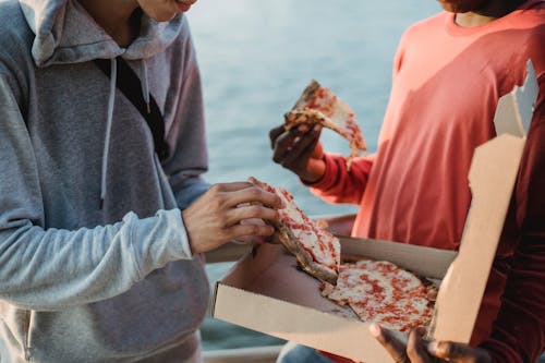 Crop anonymous male friends in casual wear eating delicious pizza together on embankment