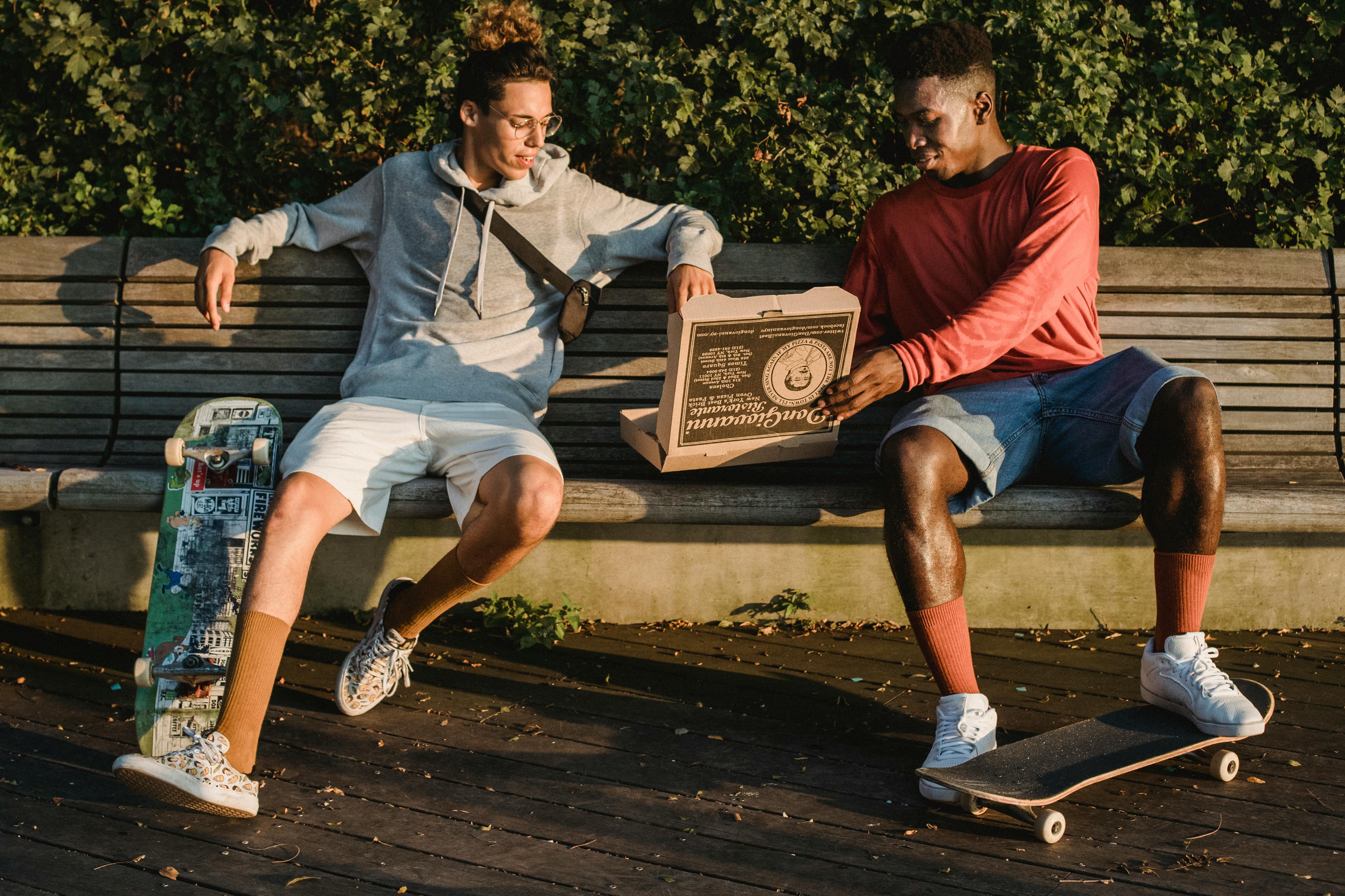 glad diverse male skaters resting on bench and eating pizza