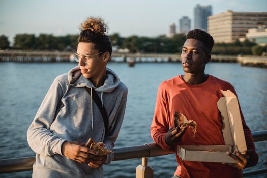 几位穿着休闲装的不同种族男性朋友，一边吃着美味的比萨饼，一边靠在城市海滨的栏杆上眺望