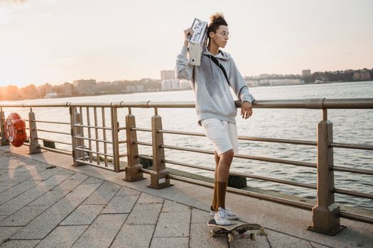 傍晚，一名长款适合种族的男性滑冰运动员肩扛老式录音机，在城市路堤上滑滑板