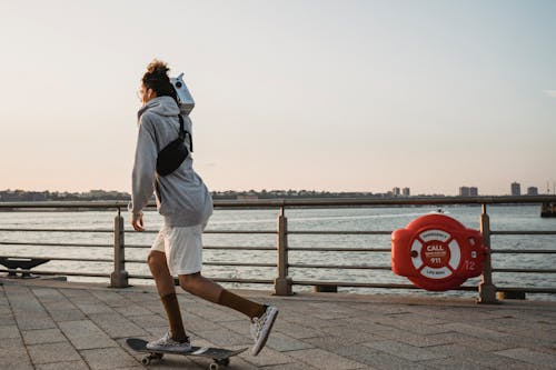 Side view full length slim male skater in casual shorts skating on city embankment at sunset