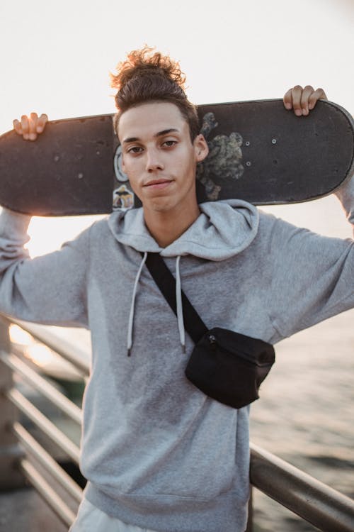 Confident young ethnic male millennial with stylish haircut in comfy hoodie standing on pier with skateboard behind head and looking at camera at sunset
