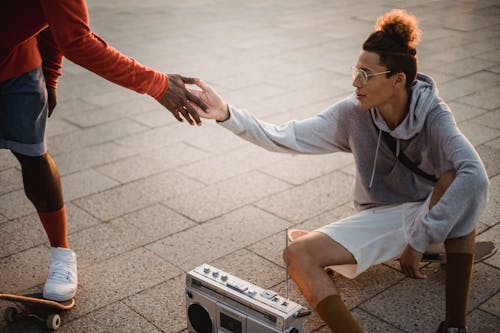 High angle of young trendy ethnic guy in stylish outfit and eyeglasses shaking hand of crop unrecognizable black friend while sitting on skateboard and listening to music with retro boombox