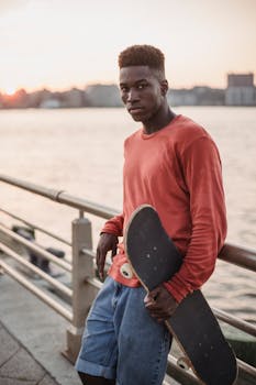 Confident young man with skateboard relaxing by the waterfront at sunset.