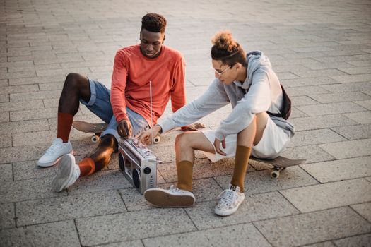 High angle of multiethnic friends listening to music on old fashioned portable sound system sitting on skateboards