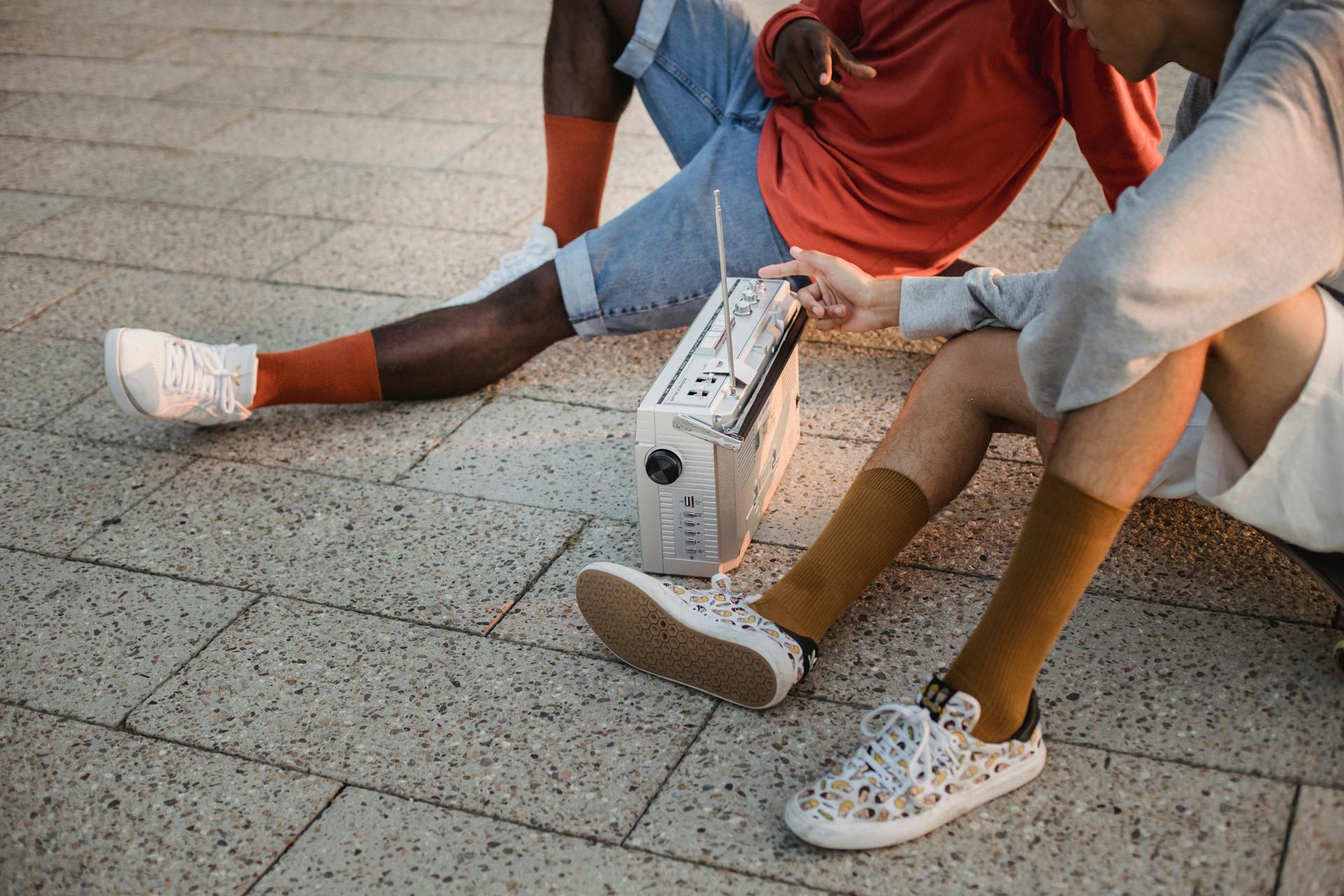 Crop multiethnic friends sitting on tiled ground and listening to music on vintage portable sound system including radio and cassette player