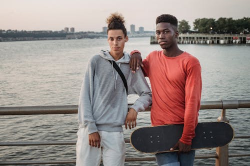 Confident multiethnic friends with skateboard leaning on fence while standing on embankment near wide river during free time together