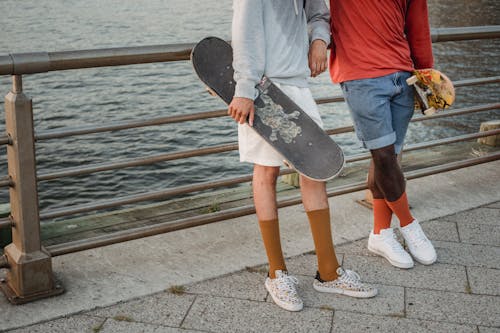 Crop anonymous male multiracial skateboarders resting on waterfront after training with skateboards together at weekend