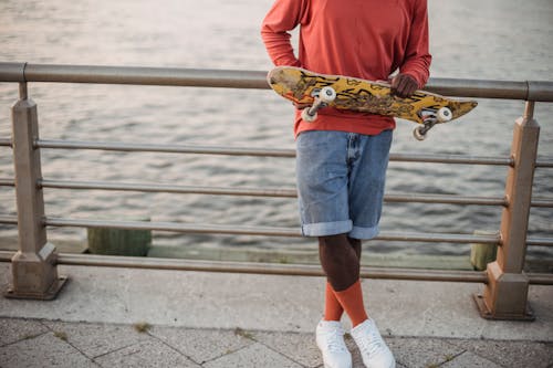 Crop anonymous African American male skateboarders with board leaning on fence standing on waterfront