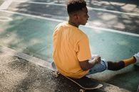 Trendy black guy sitting on skateboard after workout in sunlight