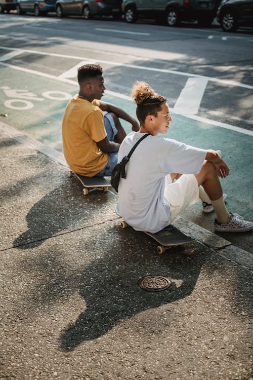 Side view of young stylish multiethnic guys in t shirts and shorts resting on asphalt road sitting on skateboards after training