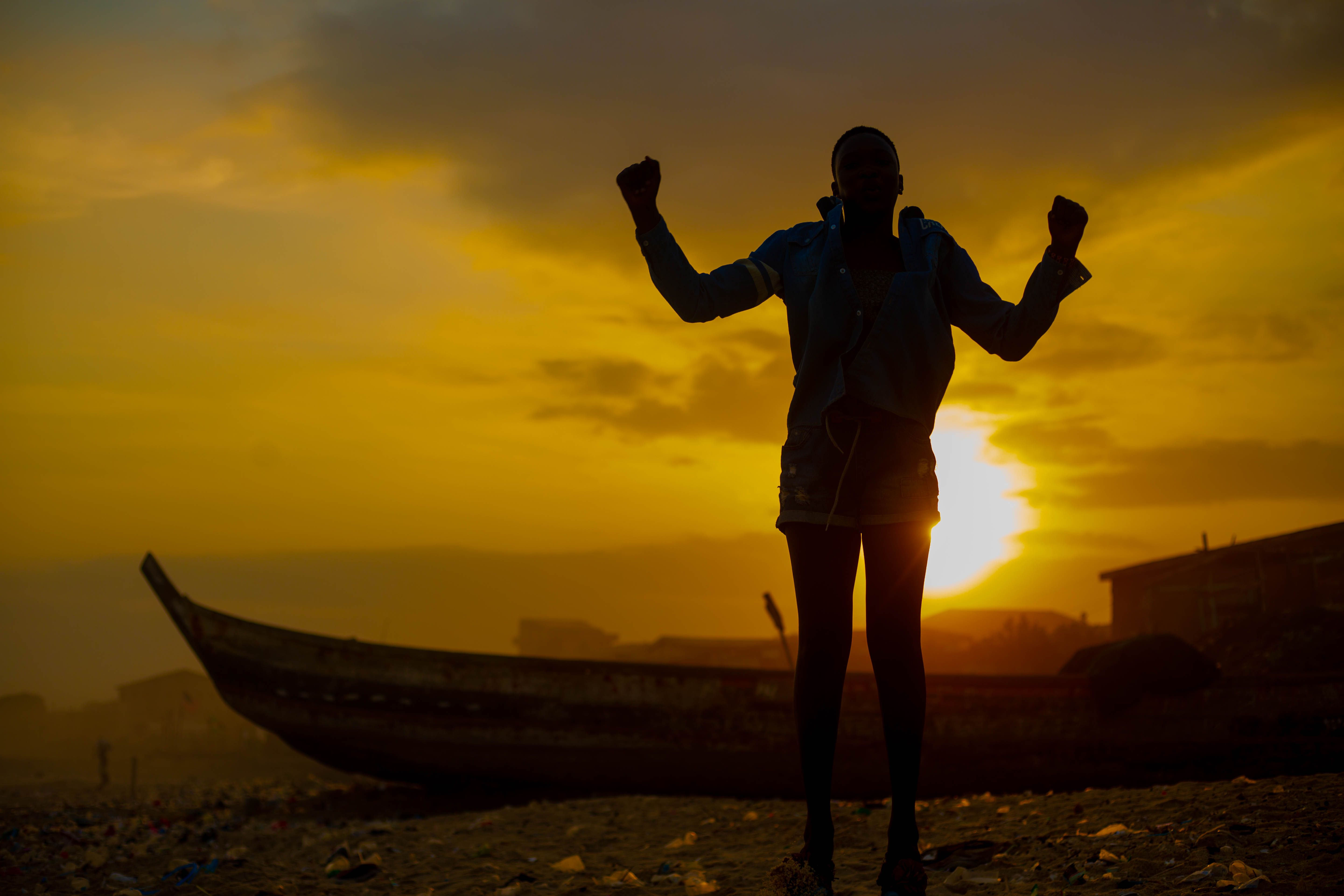 Silhouette of anonymous person standing on ground with raised hands near boat against bright sun setting on sky in evening time