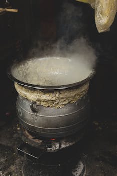 Metal cauldron with steaming water placed on metal rack with burning coal on street and emitting smoke in daylight outside