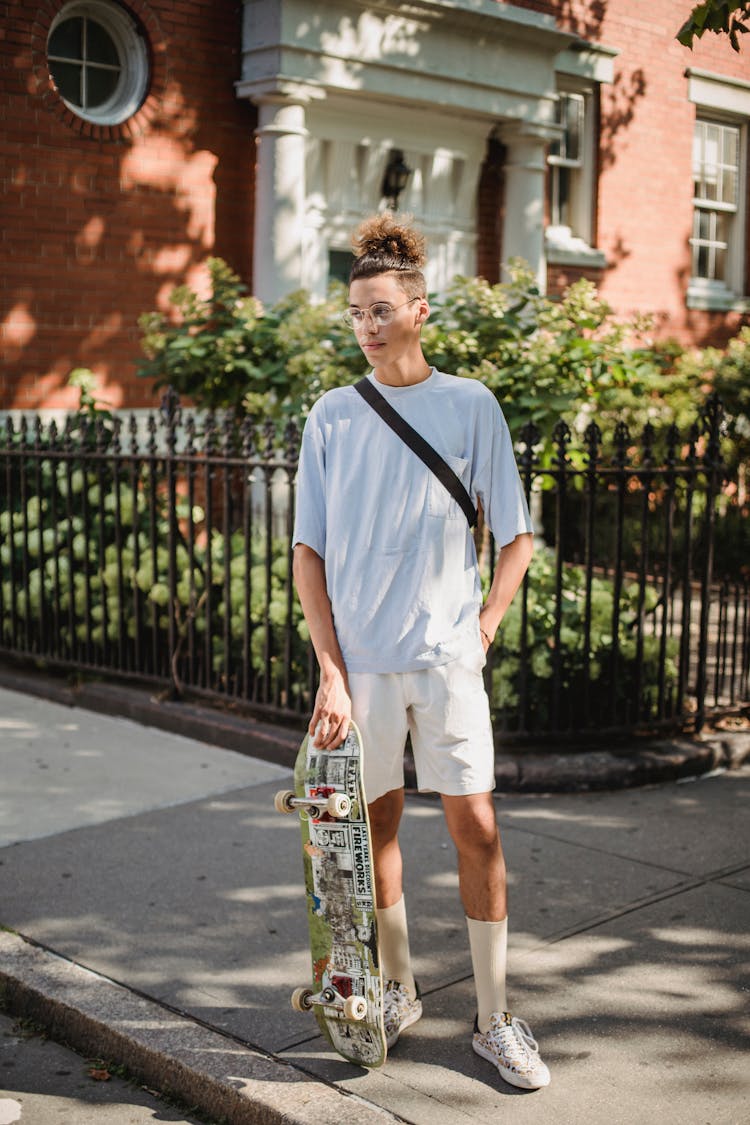 Stylish Ethnic Male Teen Relaxing At Roadside With Skateboard On Sunny Day