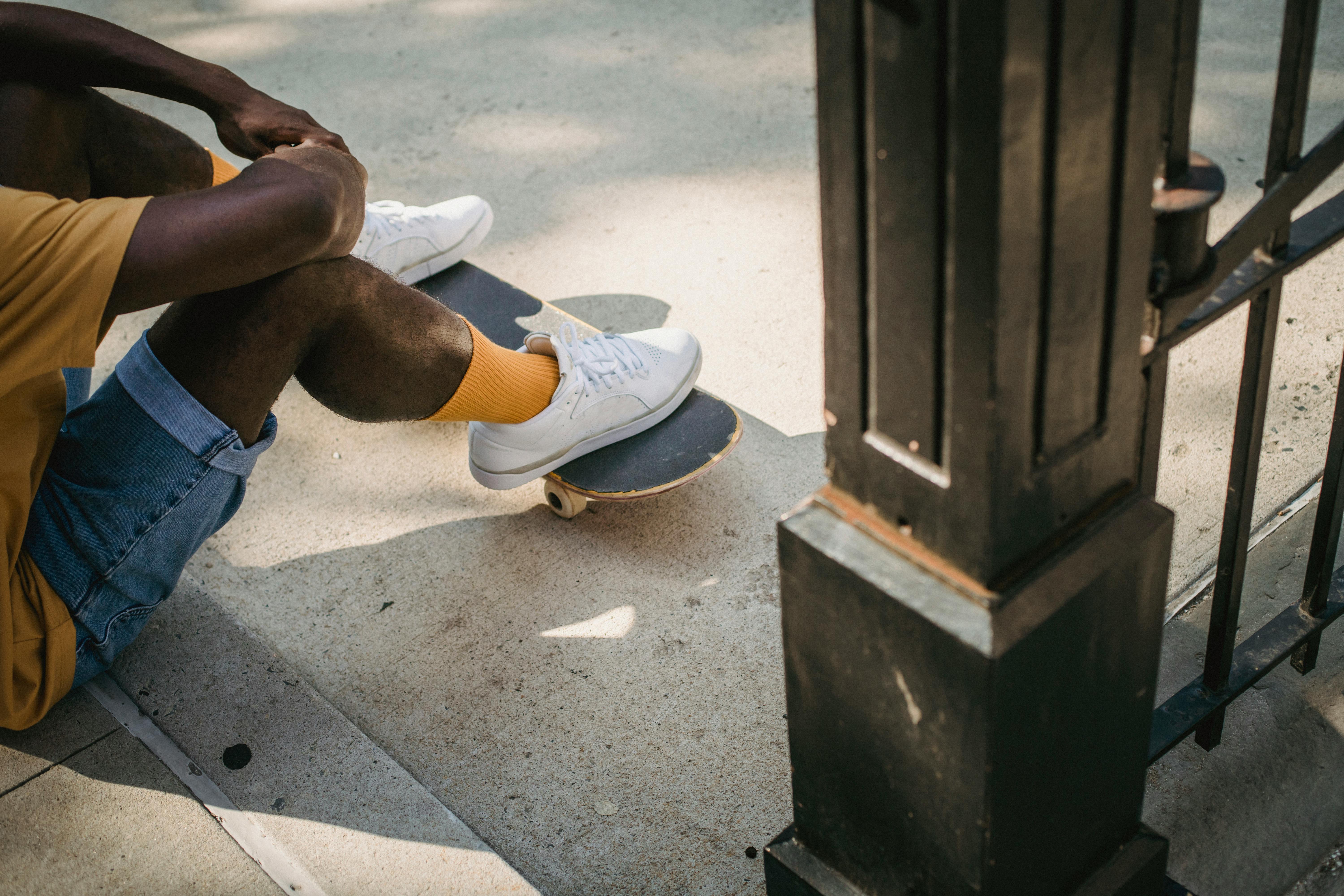 crop unrecognizable black man sitting on sidewalk with skateboard