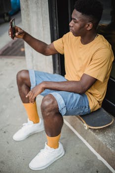 An adult man sitting on a skateboard takes a selfie outdoors, showcasing street style and relaxed vibes.