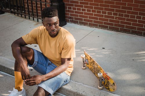 レンガ造りの建物の近くの歩道で休んでいる自信を持って若い黒人男性スケーター