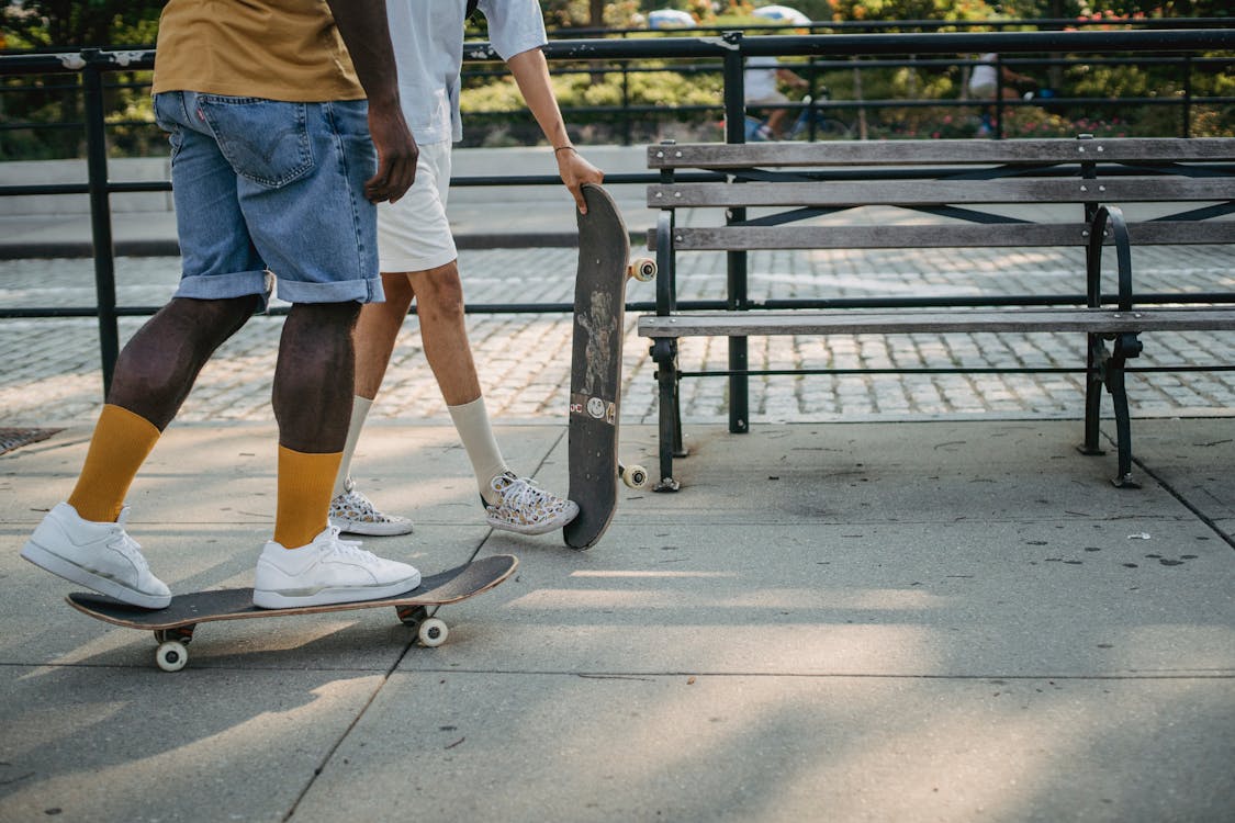 Free Crop faceless diverse skaters in casual clothes riding skateboards on asphalt walkway near fence and bench in city in sunny day Stock Photo