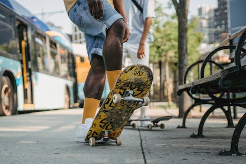 Crop unrecognizable male in yellow socks standing on colorful skateboard tail and deck at angle with friend while waiting for bus on bus stop