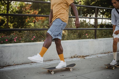 Crop anonymous black guy and male friend spending free time together while riding skateboards near park with blooming flowers on sunny day