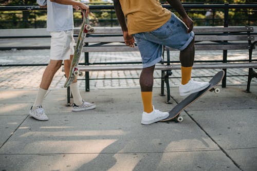 Crop unrecognizable African American guy standing on skateboard while male friend standing behind with board on city street on sunny summer day