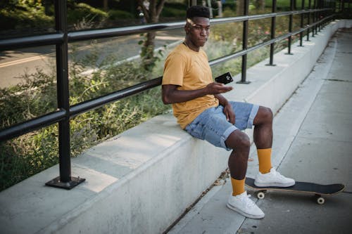 Full body of thoughtful young African american guy with skateboard wearing casual outfit sitting on concrete basement of black metal railing and throwing smartphone on sunny summer day
