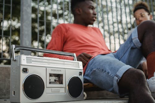 Free From below of crop young African American male friends listening to music on retro cassette recorder with radio while sitting near metal grid of park Stock Photo