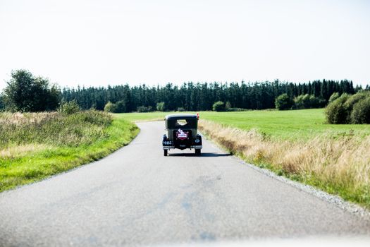 A vintage car driving on an open rural road surrounded by green fields.