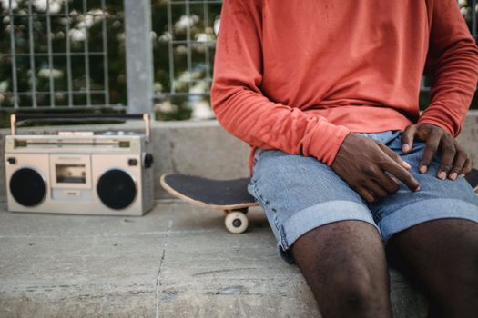 Crop anonymous African American man wearing casual clothes sitting on black skateboard near metal railing and listening to music on vintage cassette recorder