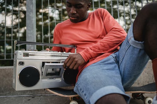 Free Crop black man in casual clothes opening retro recorder to change cassette during resting on concrete surface with skateboard in city street Stock Photo