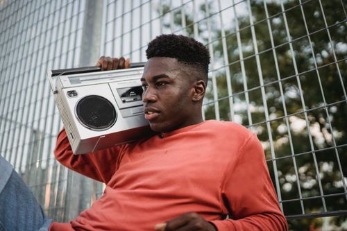 Free Young pondering African American male listening to radio from old tape recorder while looking forward near grid fence in town Stock Photo