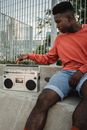 Young African American male athlete turning on old cassette recorder while sitting on skateboard near grid fence in town