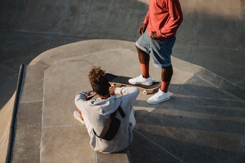 From above of crop anonymous black sportsman on skateboard standing near partner on platform in twilight