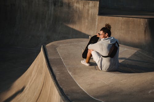 Back view of young content male athlete in casual clothes with skateboard sitting on platform while looking away in skate park