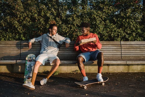 Full length of positive young multiracial friends in casual outfits sitting on bench with skateboards preparing to eat pizza