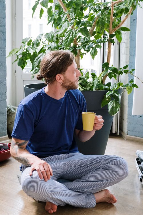 Free A man enjoying a coffee while sitting in pajamas next to houseplants indoors. Stock Photo