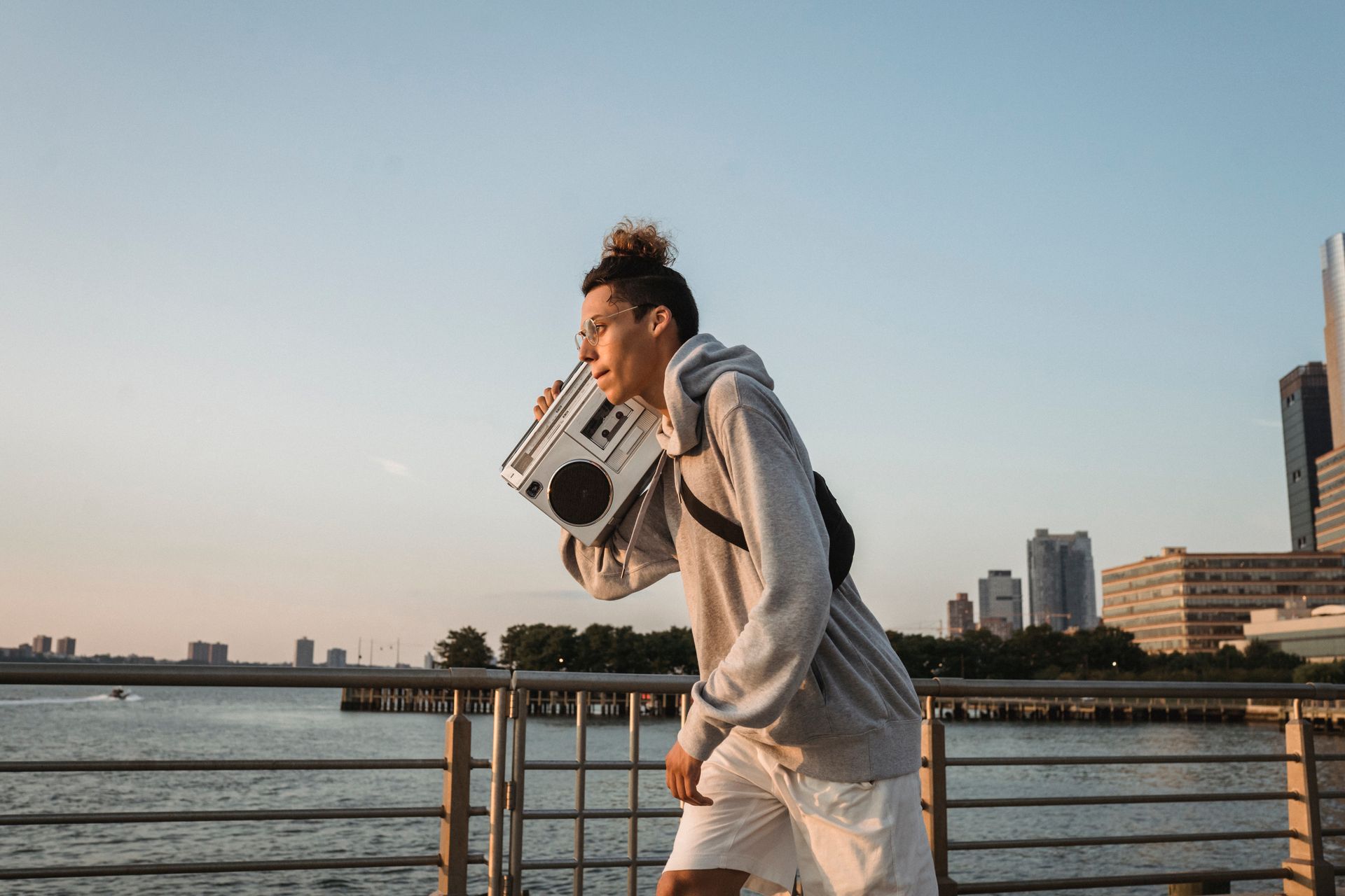 Side view of young positive male in hoodie and shorts carrying boombox while walking on embankment