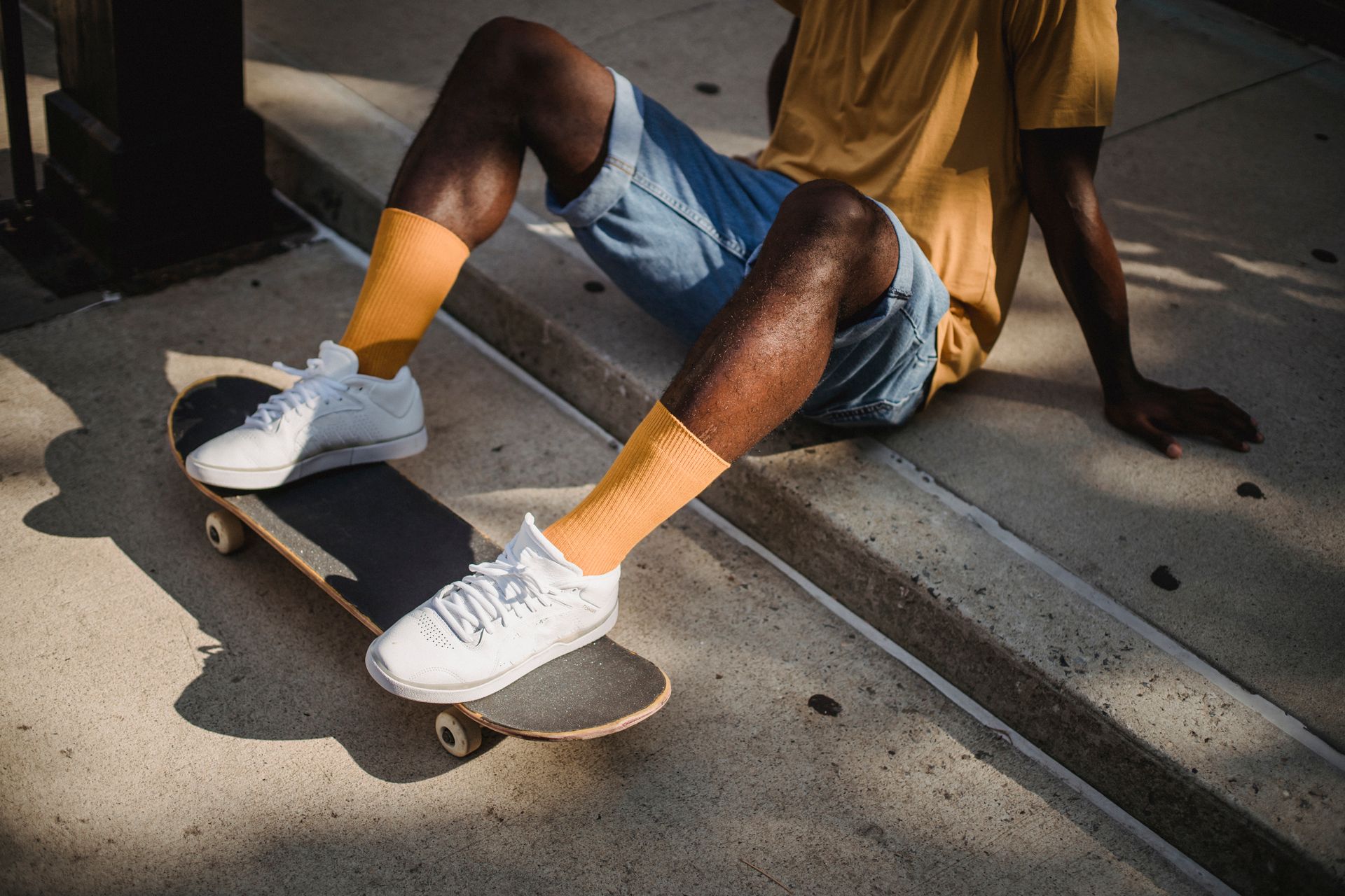 Crop anonymous African American male skater in t shirt and denim shorts sitting on sidewalk with feet on skateboard