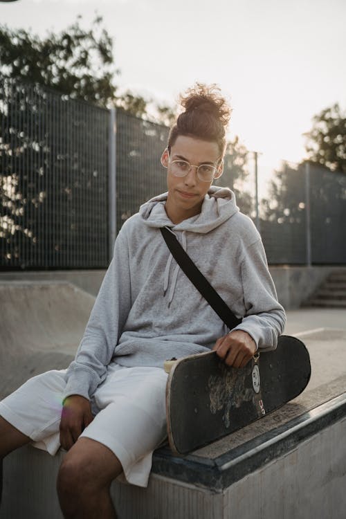 Happy young male skater in casual outfit sitting on concrete ramp with skateboard in skate park