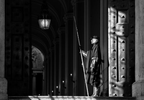 Free A Swiss Guard stands at the Vatican entrance, captured in monochrome, showcasing historic architecture. Stock Photo