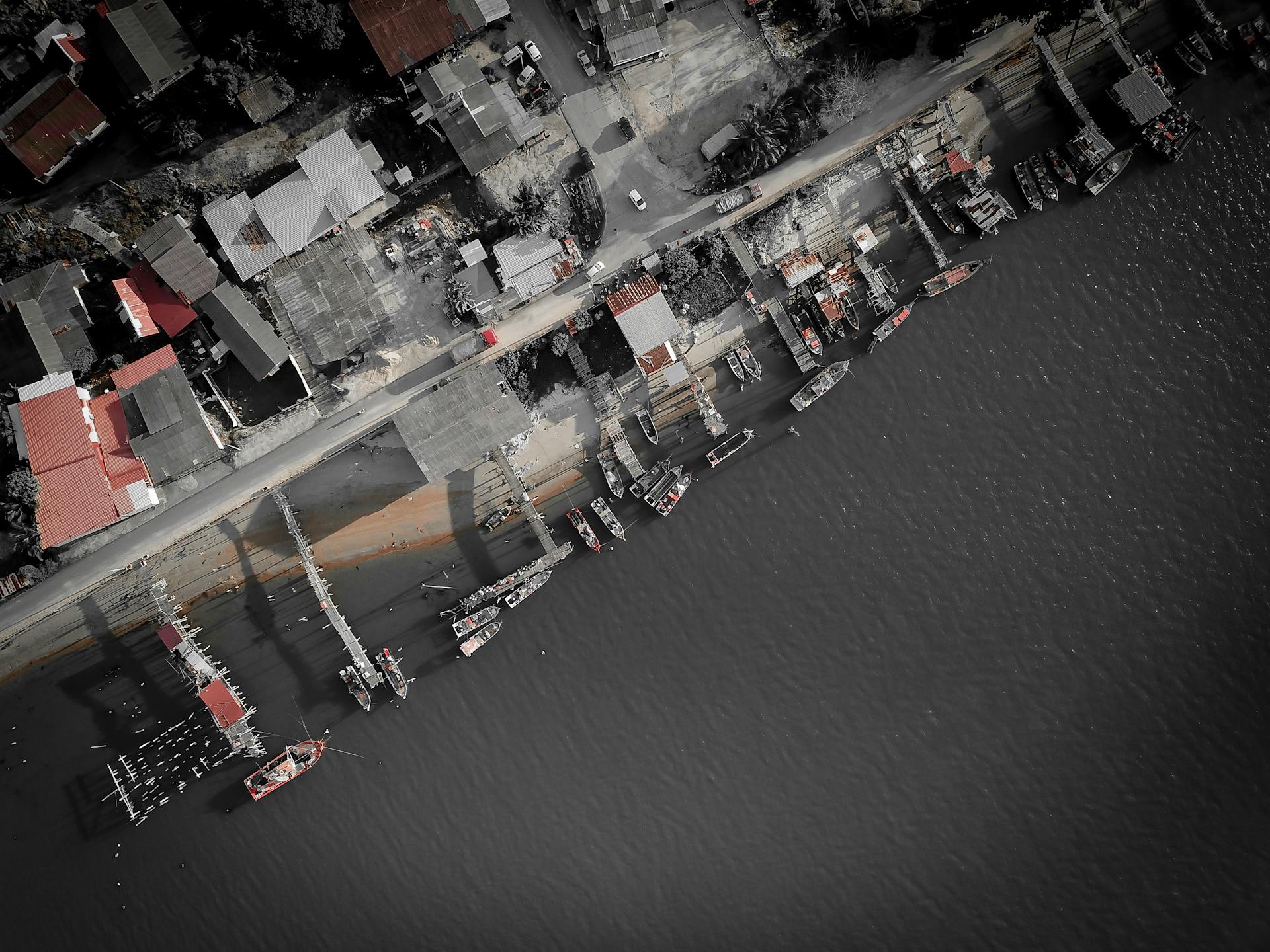 Drone view of coastal village with small houses and parked cars placed near berth with moored boats on calm sea at daytime