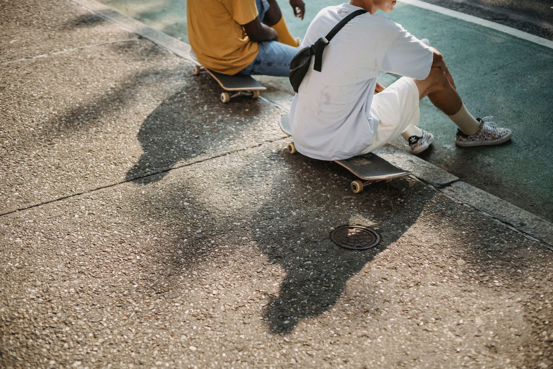 From above of crop anonymous multiethnic male friends sitting on skateboard while spending free time on asphalt road on summer sunny day