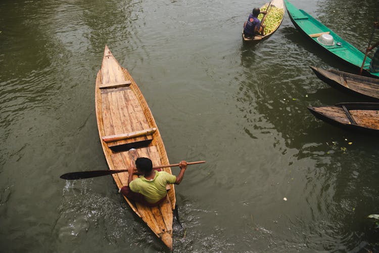 Ethnic Man Swimming On Canoe Along Boats In River