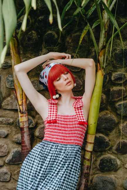 Gorgeous young female in checkered trendy dress standing with arms crossed and eyes closed in abundant tropical garden