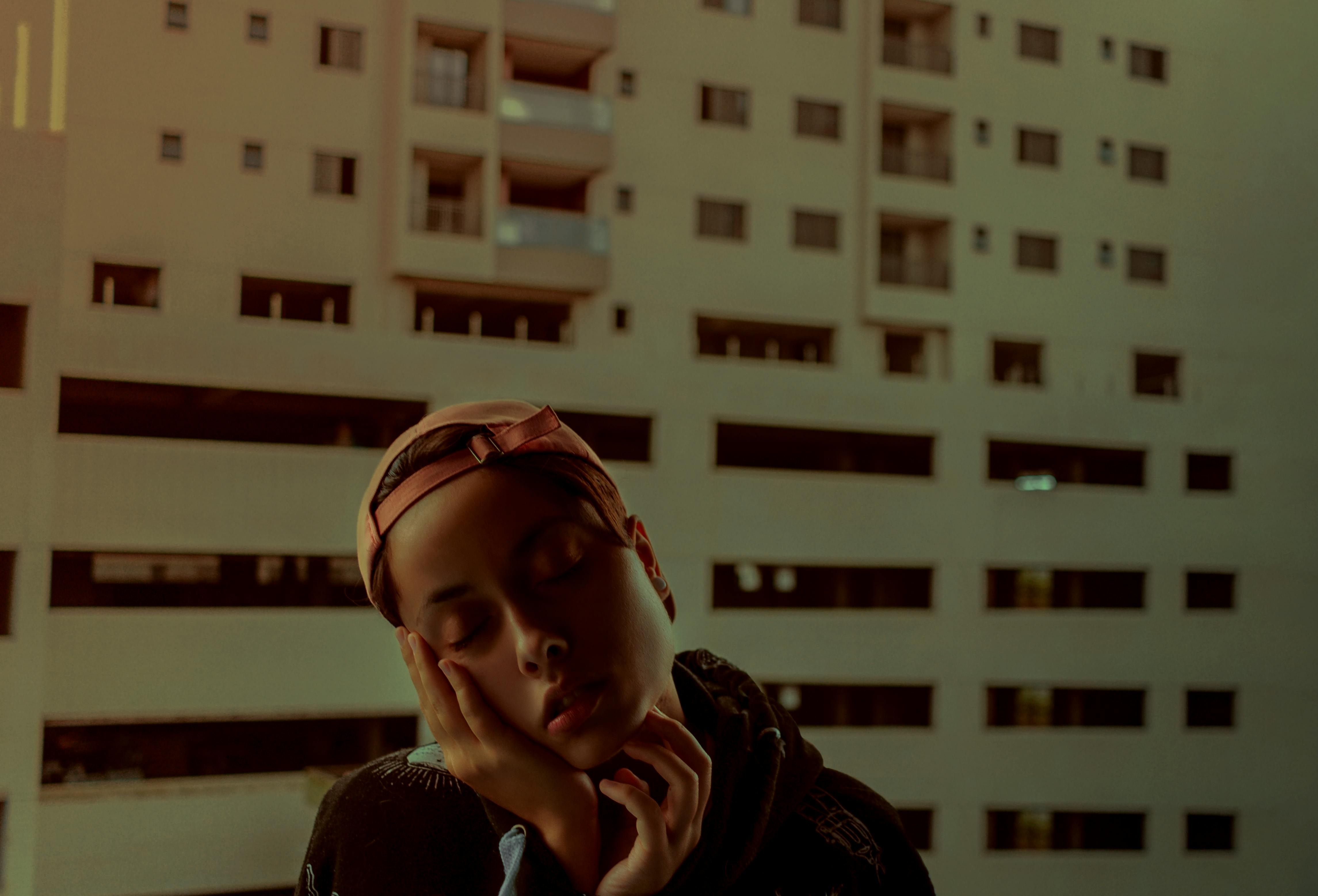 A thoughtful woman with eyes closed stands against a city building backdrop, reflecting tranquility.