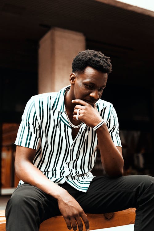 Free Thoughtful young man sitting outdoors in a striped shirt, deep in thought. Stock Photo