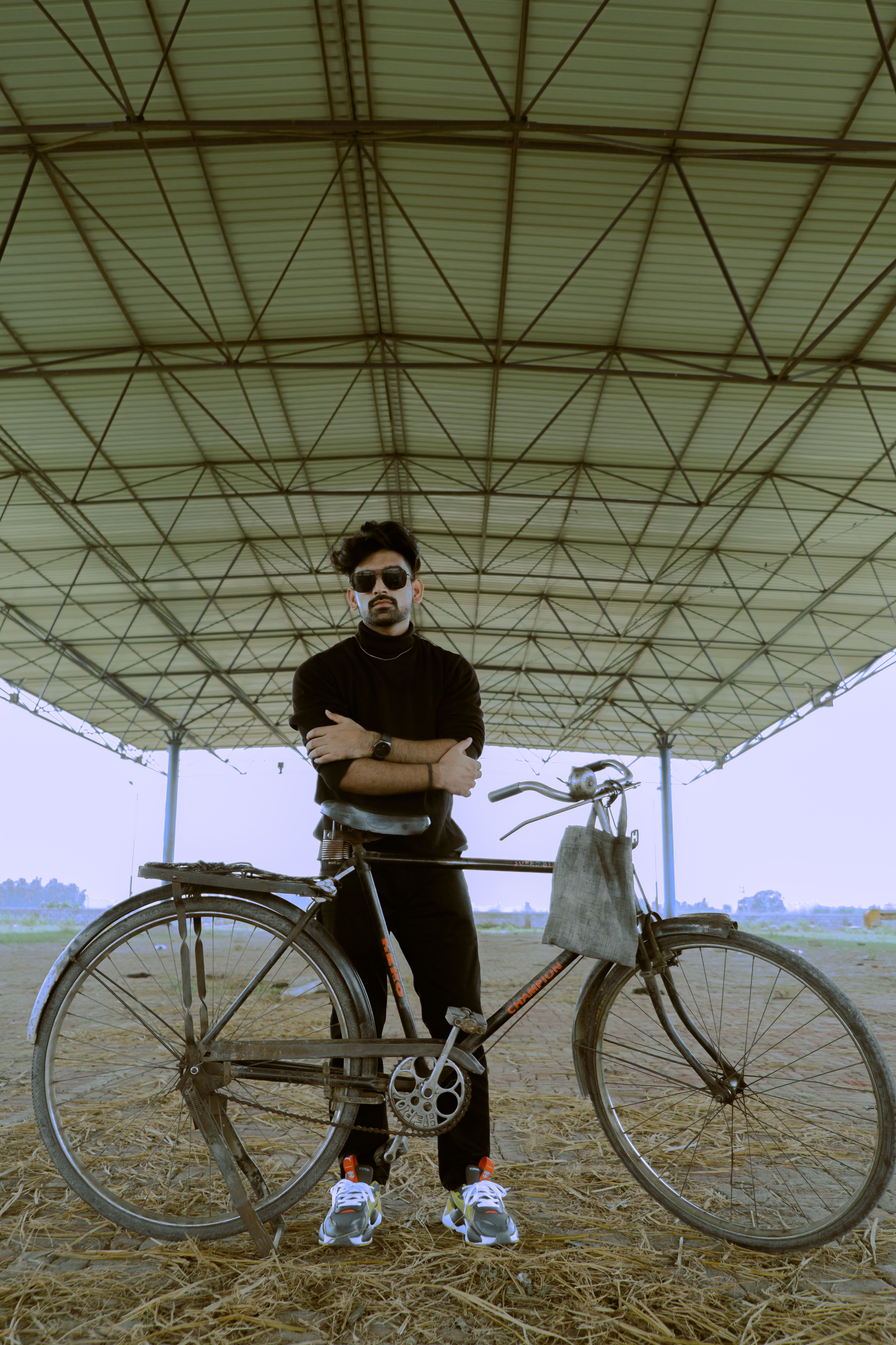 Stylish man in black stands with his bicycle under a metal canopy.