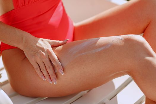 Close-up of a woman applying sunscreen on her legs wearing a red swimsuit while sunbathing.